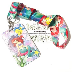 May include: A lanyard featuring a colorful design with illustrations of Ariel and Flounder from The Little Mermaid. The lanyard has a matching card holder with the same characters and a metal clasp. The text "MARIE SCOLARO" is visible.
