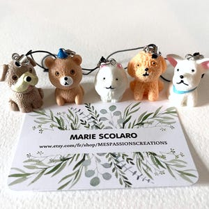 Peut inclure: Cinq petits porte-clés en forme d'animaux, colorés. Les porte-clés sont en plastique et représentent un chien brun, un chien blanc, un ours brun portant un chapeau bleu, un chien brun et blanc et un chien blanc aux oreilles roses. Les porte-clés sont disposés sur une surface blanche avec un fond vert feuillu.