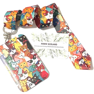 May include: A colorful lanyard and cardholder set featuring a repeating pattern of various cat illustrations. The lanyard has a silver clasp and the cardholder is filled with cat faces. A business card is also included.