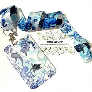 Puede incluir: Un cordón y un tarjetero a juego con un diseño de vida marina. El patrón azul y blanco incluye tortugas, delfines y estrellas de mar. El cordón tiene un cierre de metal. El tarjetero tiene una ventana de plástico transparente.