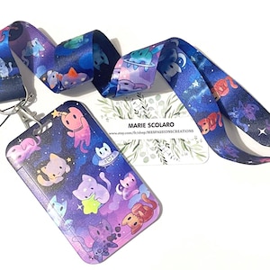 May include: A lanyard and cardholder set featuring a space cat design. The lanyard and cardholder have a dark blue background with colorful cartoon cats, stars, and planets. The cardholder has a silver metal clip and a clear plastic window.