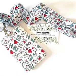 Puede incluir: Un cordón, un tarjetero y una tarjeta de visita blancos con un patrón de temática médica con corazones rojos, dientes, vendas y equipos médicos. El cordón tiene un cierre plateado. La tarjeta de visita muestra el texto "MARIE SCOLARO".