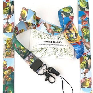 May include: A blue and green lanyard with a repeating pattern of Shrek and Fiona from the Shrek movie franchise. The lanyard has a black breakaway clasp and a black metal key ring. The lanyard is on a white background with a business card that reads "Marie Scolaro www.etsy.com/shop/MESPASSIONCREATIONS".