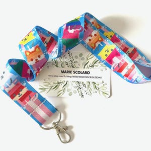 Peut inclure: Un cordon bleu avec un motif répété de chats de dessin animé de différentes couleurs. Le cordon a un fermoir argenté et une carte de visite blanche avec du texte et un motif floral.