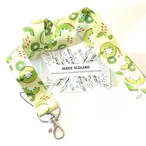 May include: A lanyard with a kiwi fruit pattern. The lanyard is light green with a repeating design of green and brown kiwi slices. It has a silver-colored metal clasp and a small tag. The business card reads "MARIE SCOLARO" and the Etsy shop URL.