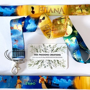 Puede incluir: Un cordón azul y verde con una imagen de Tiana de la película de Disney La Princesa y la Rana. El cordón tiene un clip plateado y una tarjeta de visita con el texto "MES PASSIONS CREATIONS" y una dirección de sitio web.