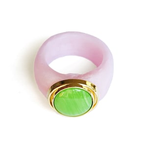 May include: A pink ring with a green stone set in a gold bezel. The ring is made of a smooth, matte material.