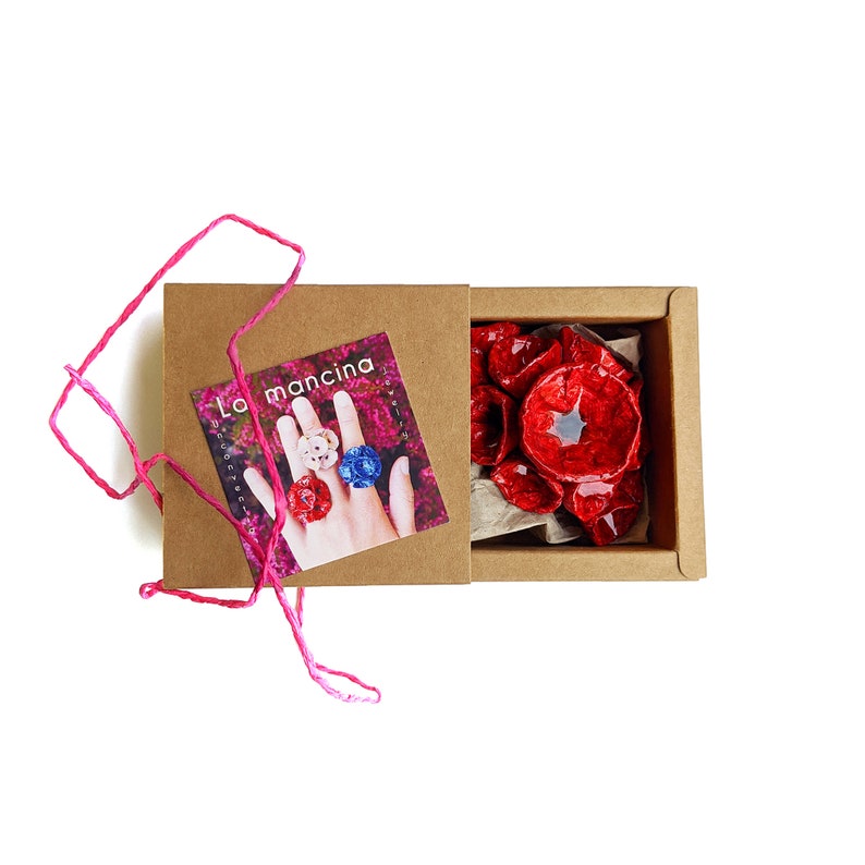 May include: A small, open box with a brown exterior and a red poppy flower design inside. The box is labelled "La mancina" and features a hand wearing three rings.