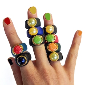 Puede incluir: Un maniquí de mano con una pila de anillos coloridos con detalles dorados y piedras preciosas de varios colores. Los anillos están hechos de arcilla polimérica negra, azul, verde y roja.