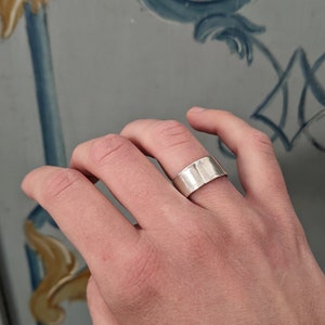 May include: A silver band ring with a hammered texture. The ring is worn on the index finger of a hand.