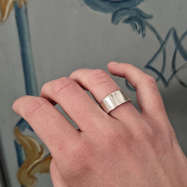May include: A silver band ring with a hammered texture. The ring is worn on the index finger of a hand.