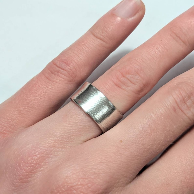 May include: A silver band ring with a hammered finish. The ring is wide and flat, and it has a simple, modern design.