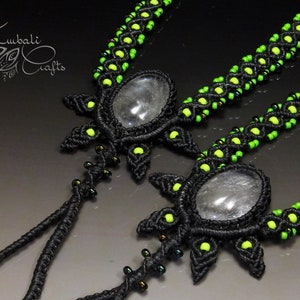 以下が含まれることがあります： 緑色のビーズと透明な水晶のペンダントが付いた、黒いマクラメのネックレスが2本。ネックレスは装飾的なパターンで結ばれており、水晶の石の周りに星形のデザインが施されています。