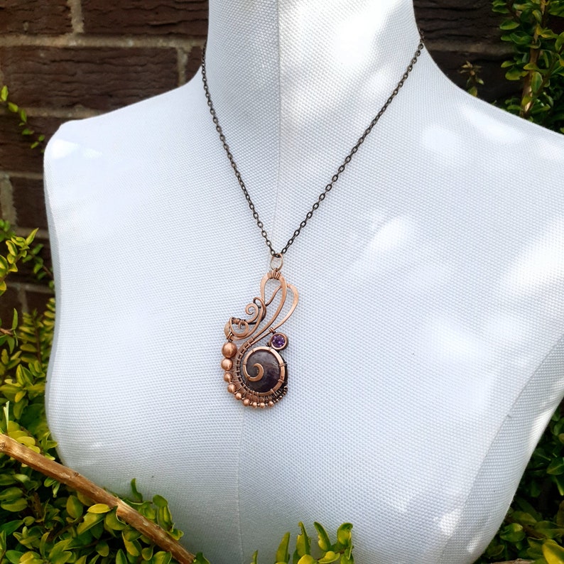 Peut inclure: Un collier en fil de cuivre avec un pendentif en pierre violette. Le pendentif est un design en spirale avec du fil de cuivre enroul&eacute; autour de la pierre. Le collier est sur un mannequin blanc.