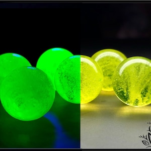 Op de afbeelding: Een verzameling bolvormige glazen objecten getoond in twee belichtingsstanden. Links lichten de bollen op met een fel neon-groen licht in het donker. Rechts verschijnen dezelfde bollen als doorschijnend geel glas met interne luchtbellen.