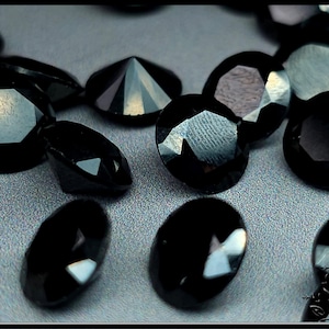 May include: A collection of loose, faceted black gemstones with a brilliant cut. Each stone features multiple polished surfaces that reflect light, creating sharp geometric patterns across the dark, opaque faces of the round and oval-shaped gems.