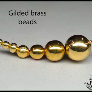 May include: Close-up of gilded brass beads in varying sizes, strung on a copper wire. The beads are a shiny gold colour, reflecting light. Text in the image reads "Gilded brass beads".