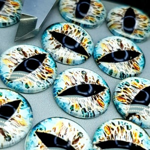 May include: A collection of round glass cabochons featuring a realistic dragon eye design with a vertical black pupil, surrounded by a textured iris in shades of blue, white, and gold, positioned next to a metal craft tool edge.