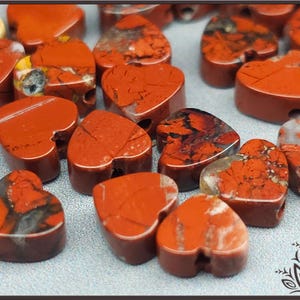 Puede incluir: Un conjunto de cuentas en forma de corazón en tonos rojos y marrones. Las cuentas tienen un acabado pulido y patrones naturales visibles. Cada cuenta tiene un pequeño agujero para ensartar. Adecuado para la fabricación de joyas.