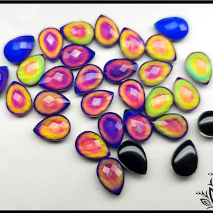 Op de afbeelding: Een verzameling druppelvormige cabochons met een gefacetteerd oppervlak. De stenen vertonen een levendig verloop van geel, oranje, roze en paars, terwijl andere effen blauw of zwart zijn, gerangschikt in een verspreid patroon op een witte ondergrond.