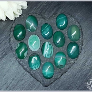 Könnte beinhalten: Eine Sammlung von 12 grünen Achat-Cabochons, jeder mit einem einzigartigen Muster aus weißen und grünen Streifen, angeordnet in Herzform auf einer dunkelgrauen Schieferfläche.