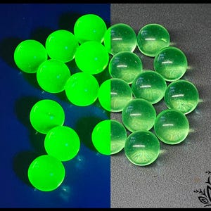 Puede incluir: Una colección de pequeñas cuentas de vidrio esféricas dispuestas en grupo. El lado izquierdo de la imagen muestra las cuentas brillando con una luz verde neón brillante, mientras que el lado derecho muestra las mismas cuentas en un color verde pálido.