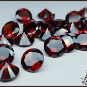 May include: A collection of loose, round-cut dark red gemstones with faceted surfaces. The stones exhibit a deep crimson hue and reflective properties, arranged in a scattered cluster on a plain, light-colored surface.