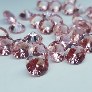 May include: A collection of round, faceted, light pink gemstones with a brilliant cut. Each stone features multiple triangular facets that reflect light, creating a sparkling appearance across the surface of these small, loose, circular crystals.
