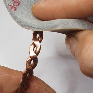 May include: A hand holding a copper braided bracelet and sanding it with a grey sanding block.
