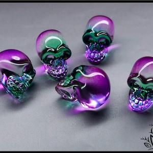 May include: Five small, translucent glass skulls arranged in a group. Each skull features a gradient colour transition from deep purple at the top to a vibrant green at the jaw and teeth area. The glass material is smooth, reflective, and polished.