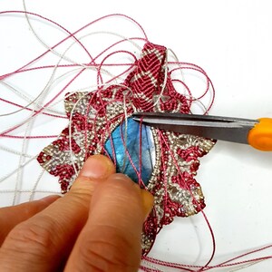 Op de afbeelding: Een close-up van een hand die een schaar vasthoudt en een draad aan een roze en witte macram&eacute; ster met een blauwe edelsteen in het midden knipt.