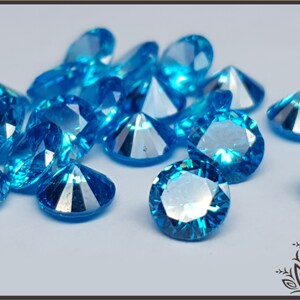 May include: A collection of loose, round-cut gemstones in a vibrant, bright blue color. Each stone features multiple facets that reflect light, creating a sparkling appearance. The stones are scattered across a plain, light-colored surface.