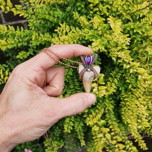K&ouml;nnte beinhalten: Ein Anh&auml;nger aus Kupferdraht, der ein zahnf&ouml;rmiges Knochenst&uuml;ck mit einem violetten Edelstein in einer Krone aus Kupferdraht umh&uuml;llt.