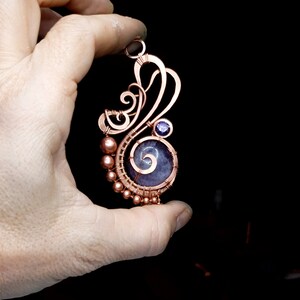 Peut inclure: Un pendentif en fil de cuivre enroul&eacute; avec une pierre violette et grise avec un motif en spirale. Le pendentif est tenu dans une main.