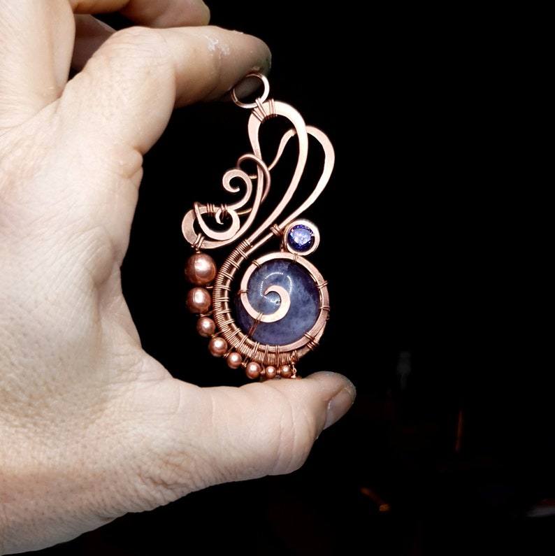 Peut inclure: Un pendentif en fil de cuivre enroul&eacute; avec une pierre violette et grise avec un motif en spirale. Le pendentif est tenu dans une main.