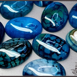 May include: A collection of oval-shaped blue agate cabochons featuring a distinct crackled pattern. Each polished stone displays varying shades of deep blue, teal, and navy with dark web-like veining throughout the smooth, glossy surfaces.