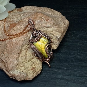 May include: A pendant featuring a yellow and green labradorite stone encased in intricate, hand-woven copper wire wrapping. The pendant hangs from a matching copper ball chain, showcasing detailed metalwork with swirls and textured wire accents.