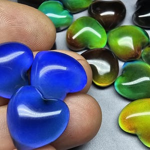 Puede incluir: Una colección de piedras de vidrio en forma de corazón iridiscentes en tonos azules, verdes y marrones. Las piedras están dispuestas sobre una mano.