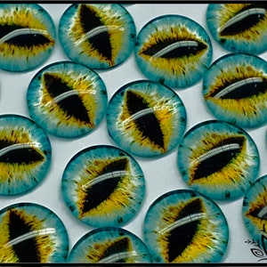 May include: A collection of circular glass cabochons featuring a realistic dragon eye design. Each piece displays a vertical black pupil surrounded by a vibrant yellow iris and a bright blue outer ring, creating a detailed reptilian appearance.