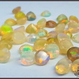 May include: A collection of round, faceted opals displaying a translucent, milky appearance with iridescent flashes of green, orange, and yellow. These small, polished gemstones are scattered across a plain surface, showcasing their reflective surfaces.