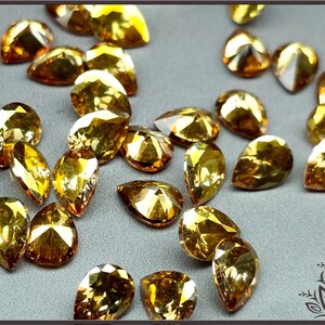 May include: A collection of loose, pear-shaped faceted gemstones in a vibrant golden-yellow hue. Each stone features multiple triangular facets that catch the light, creating a sparkling effect across the scattered arrangement of these small, teardrop-shaped jewels.
