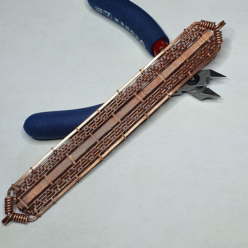 K&ouml;nnte beinhalten: Ein Armband aus Kupferdraht mit geometrischem Muster. Das Armband wird von einer blauen Zange gehalten.