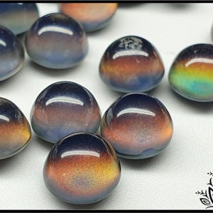 Op de afbeelding: Een verzameling iriserende glazen cabochons in tinten blauw, paars en goud. De cabochons zijn rond en hebben een glad, gepolijst oppervlak.