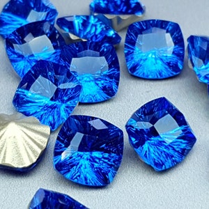 Puede incluir: Un grupo de gemas de vidrio azul, facetadas, de forma cuadrada con un patrón de estrella. Las gemas están dispersas sobre una superficie gris.