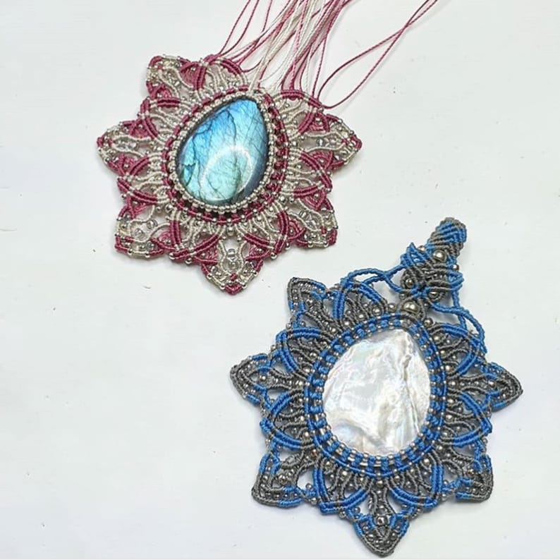 Op de afbeelding: Twee handgemaakte macram&eacute; hangers met edelsteen accenten. De bovenste hanger heeft een blauwe labradorite steen in een zilveren setting, omringd door een rood en wit macram&eacute; ontwerp. De onderste hanger heeft een witte iriserende schelp in een zilveren setting, omringd door een blauw en zilver macram&eacute; ontwerp.