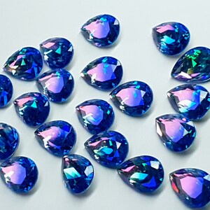 May include: A collection of iridescent blue teardrop-shaped glass rhinestones. The rhinestones have a holographic finish that shimmers with different colors in the light.