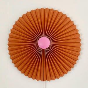May include: A large, round, brown paper fan with a pink circle in the center. The fan is folded into many pleats, creating a radial pattern.