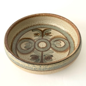 May include: A round, ceramic bowl with a decorative design. The bowl features a beige interior with a central pattern of brown and gray concentric circles and stylized shapes. The rim is a darker brown.