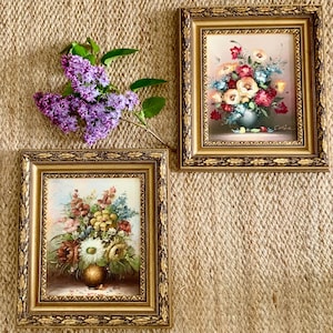 May include: Two framed floral paintings with gold ornate frames. The paintings depict bouquets of colourful flowers in vases. A sprig of purple lilacs rests on the textured background. The paintings are a vintage style.