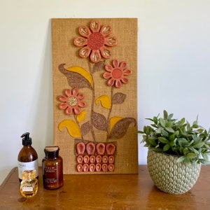 May include: A brown burlap wall hanging featuring a 3D floral design. The flowers are crafted from felt and have a red, orange, and yellow colour scheme. The flowers are arranged in a vase shape and are surrounded by green leaves.